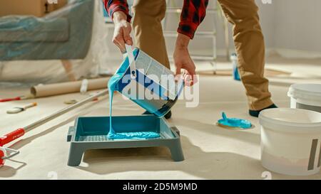 Jeune homme en Jean marron et en chemise à carreaux rouges pique un pot de peinture bleu clair et le verse dans un plateau. Rénovation des chambres à la maison. Banque D'Images