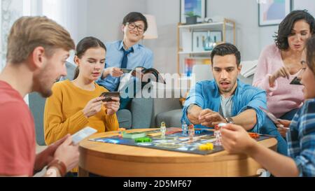 Groupe diversifié de gars et de filles jouant dans un jeu de plateau stratégique avec cartes et Dice. Salon confortable en journée Banque D'Images