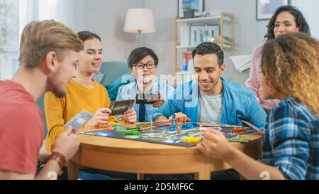 Groupe diversifié de gars et de filles jouant dans un jeu de plateau stratégique avec cartes et Dice. Salon confortable en journée Banque D'Images