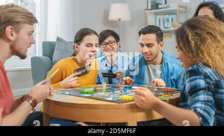 Groupe diversifié de gars et de filles jouant dans un jeu de plateau stratégique avec cartes et Dice. Salon confortable en journée Banque D'Images