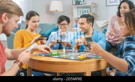 Groupe diversifié de gars et de filles jouant dans un jeu de plateau stratégique avec cartes et Dice. Salon confortable en journée Banque D'Images