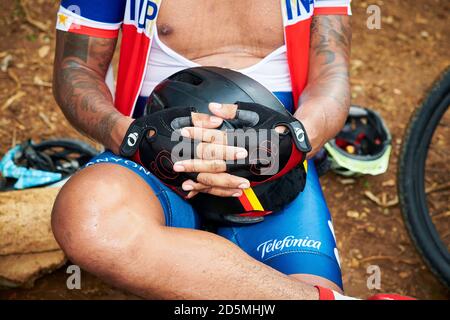 Vue rapprochée d'un cycliste tatoué dans une tenue de course repose et porte son casque avec ses mains, portant des gants de vélo Banque D'Images