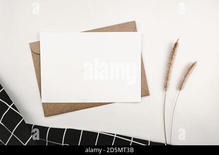 Papier blanc vierge sur enveloppe en papier marron avec fleur sèche de queue d'éponge et tissu noir avec motif de grille blanc. Maquette de gre vide horizontal Banque D'Images