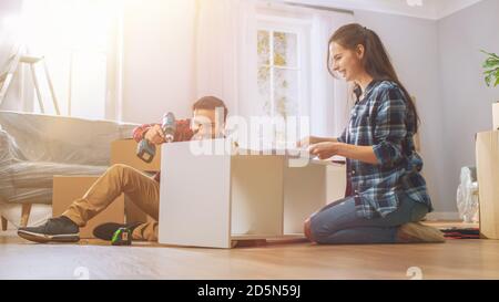 Le couple heureux assemble le mobilier en équipe, la fille lit les instructions et le garçon serre la vis avec une perceuse. Déménagement dans un nouvel appartement, couple Assemble Banque D'Images