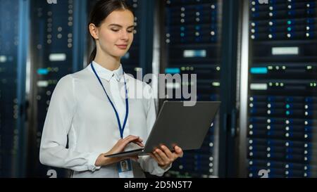 Magnifique technicien INFORMATIQUE féminin de centre de données situé près du couloir du rack de serveurs avec un ordinateur portable. Elle utilise un ordinateur portable et travaille avec des serveurs. Banque D'Images