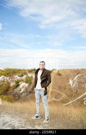 Homme sérieux avec des lunettes en jeans, t-shirt et cape se tient dans un terrain vallonné. Un homme adulte avec des lunettes dans la capuche a une vue très bien loin dans la campagne Banque D'Images