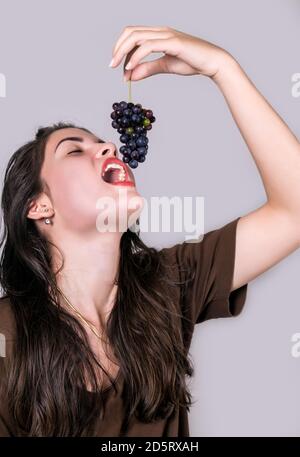 Jeune fille sur le point de manger un bouquet de petits raisins mûrs qui sont sur le point d'entrer dans sa bouche ouverte, elle tient la tige du bouquet de raisins Banque D'Images