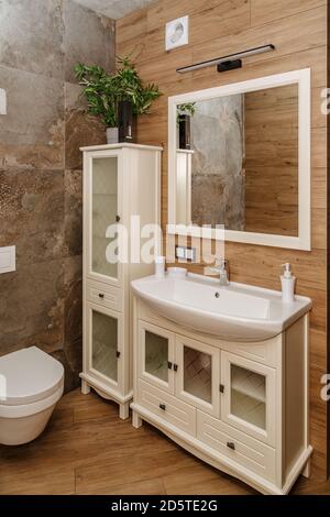 mobilier de salle de bains blanc dans un style moderne, distributeur d'eau blanc, miroir carré et une partie des toilettes intégrées sur un fond de carrelage en bois Banque D'Images