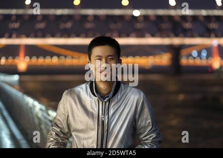 Prise de vue moyenne d'un jeune homme asiatique regardant l'appareil photo la nuit. Sourire heureux. Le pont du fleuve Yangtze de Wuhan a été défoqué comme arrière-plan Banque D'Images