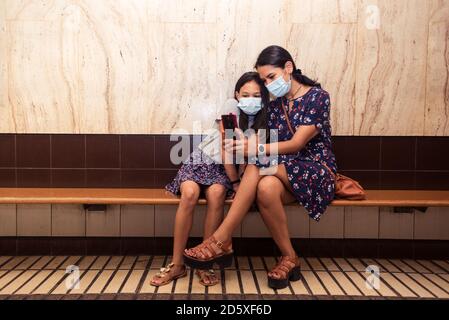 Petite fille et sa jeune mère prenant un selfie portant un masque chirurgical. Les filles sont assises sur le banc à la gare. Banque D'Images