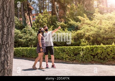 Couple marchant le jour ensoleillé dans le parc. Homme pointant avec la main. Jeune couple portant un masque de protection. Banque D'Images