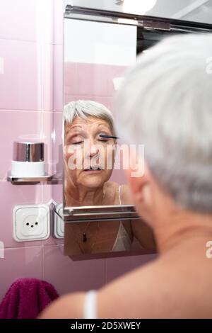 Femme caucasienne âgée peignant ses yeux dans son miroir de maison Banque D'Images