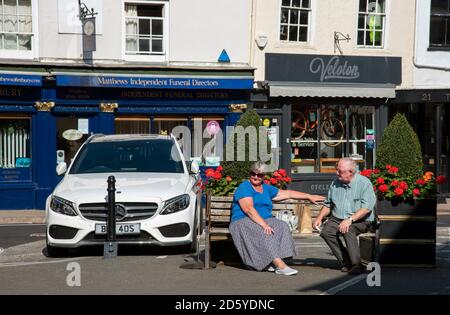 Tetbury, Gloucestershire, Angleterre, Royaume-Uni. 2020. Couple de personnes âgées assis sur un banc et un quartier commerçant de détail dans le centre-ville de Tetbury, Gloucestershir Banque D'Images