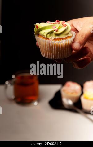 La main d'un homme tient un délicieux cupcake sur le fond d'une assiette avec plusieurs cupcakes colorés et thé chaud. Aliments malsains et sucrés Banque D'Images