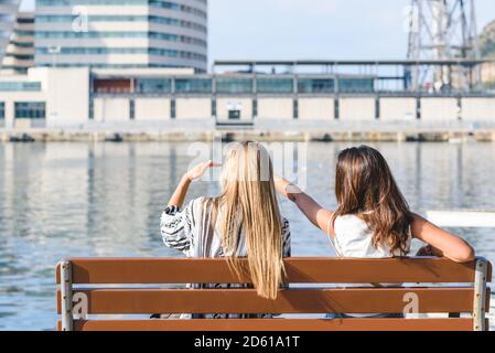 vue arrière de deux amies assises sur un banc et en pointant quelque chose qui saisit leur intérêt pour un port zone Banque D'Images