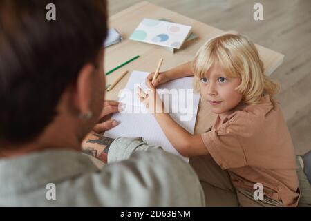 Petit fils aux cheveux blonds parlant à son père pendant assis à la table et dessin Banque D'Images