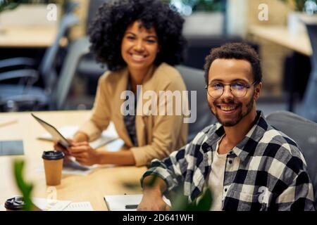 Deux jeunes employés de bureau gaies, hommes et femmes, assis au bureau, souriant à la caméra tout en travaillant ensemble dans un espace de travail de collègue. La vie de bureau, le travail d'équipe et les affaires Banque D'Images