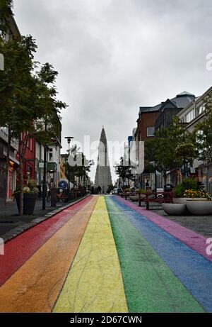 Une route peinte en arc-en-ciel (Skolavordustigur) mène à l'église Hallgrimskirkja à Reykjavik, en Islande. Des arbres et des plantes bordent la rue remplie par le magasin. Banque D'Images