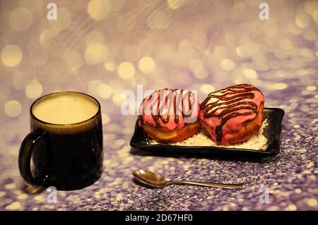 Tasse à cappuccino en verre avec mousse et assiette rectangulaire noire avec deux beignets en glaçage rose avec chocolat et poudre sur fond gris. Gros plan. Banque D'Images