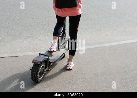 Les jambes d'une jeune fille inconnue en baskets blanches et leggings de gym sur un scooter électrique noir au-dessus de l'asphalte urbain. Transport moderne, électrique Banque D'Images