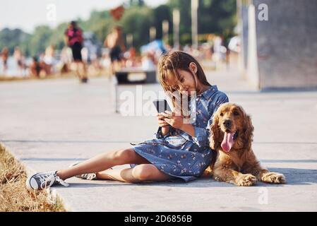 Se trouve sur le sol. Avec un smartphone entre vos mains. Petite fille mignonne ont une promenade avec son chien à l'extérieur à la journée ensoleillée Banque D'Images