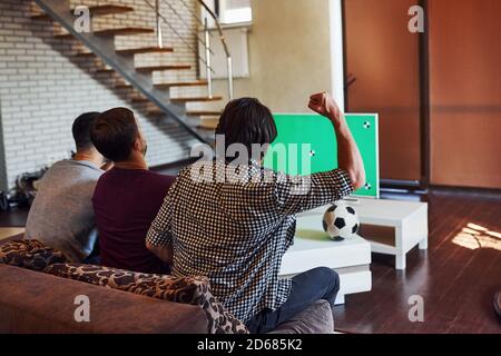 Avec les mains levées. Trois amis enthousiastes regardant le football à la télévision à la maison ensemble Banque D'Images