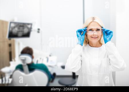 Portrait d'une femme dentiste souriant et regardant la caméra, clinique de dentisterie de fond avec un patient. Lunettes de protection réglables par un dentiste Banque D'Images
