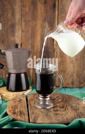 Versez le lait dans le café. Faire du café avec de la crème. Tête en verre à la main. Un verre de cappuccino. Machine à café Geyser sur une table en bois. Faites cuire le petit déjeuner à Banque D'Images