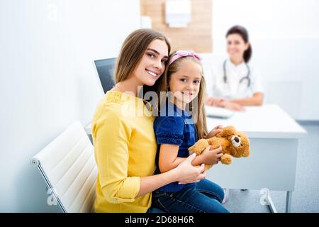 Portrait d'une petite fille heureuse assise dans les bras de sa mère à l'hôpital. Pédiatre en visite avec un parent, santé des enfants Banque D'Images