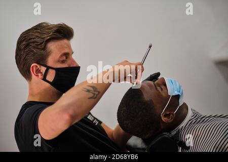 Un jeune coiffeur tatoué coupe les cheveux d'un client tout en portant Un masque facial conforme à la réglementation COVID-19 Banque D'Images