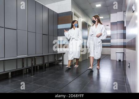 Jeunes filles du corps entier dans des peignoirs doux et des masques pour le visage parler et marcher près des casiers dans le salon de spa Banque D'Images