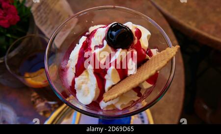 Vue rapprochée d'une délicieuse coupe de sundae à la crème glacée avec sauce rouge, crème fouettée, gaufres et cerise sur une table de café avec un verre de soda. Banque D'Images