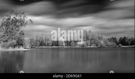 Exposition longue en noir et blanc d'une vue sur un lac vers une île avec une forêt de style scandinave de des bouleaux et une cabane en rondins Banque D'Images