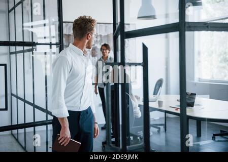 Regarde derrière. Homme dans des lunettes avec bloc-notes dans les mains se tient dans le bureau en face des employés Banque D'Images