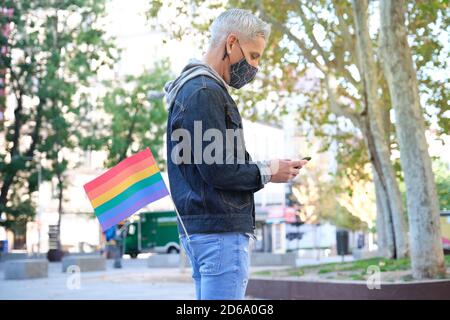 Jeune homosexuel caucasien portant un masque facial avec un drapeau LGBT dans sa poche discutant sur son smartphone. Célébration de la fierté LGBT en temps de pandémie. Banque D'Images
