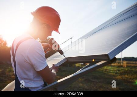 À l'aide d'un tournevis sans fil. Homme travaillant en uniforme bleu à l'extérieur avec des piles solaires à la journée ensoleillée Banque D'Images