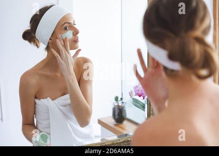 Femme appliquant le paquet de visage tout en regardant dans le miroir dans salle de bains Banque D'Images