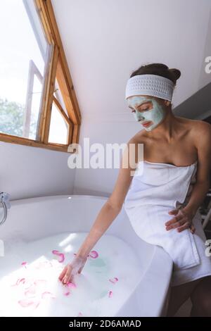 Femme avec masque tombant pétales de rose tout en étant assise bord de la baignoire dans la salle de bains Banque D'Images