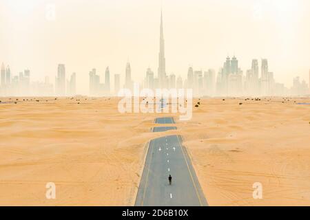 Vue aérienne d'une personne non identifiée marchant sur une route déserte recouverte de dunes de sable avec la ligne d'horizon de Dubaï en arrière-plan. Banque D'Images