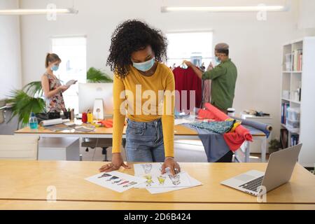 Femme designer de mode portant un masque de visage regardant les croquis de conception de vêtements en studio Banque D'Images