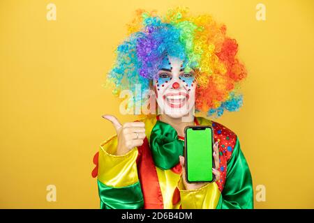 Clown debout sur fond jaune insolé jaune souriant, montrant le téléphone et pointant sur le côté Banque D'Images