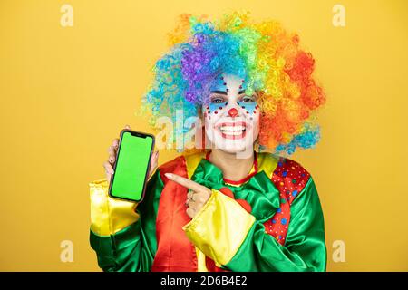 Clown se tenant sur fond jaune insolé jaune souriant, montrant et pointant le téléphone Banque D'Images