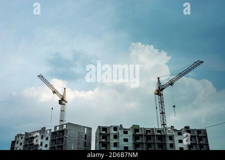 Grues au-dessus de panneaux gris non finis à plusieurs étages sur fond de ciel bleu ciel nuageux avec espace de copie. Processus de construction de l'immeuble d'appartements Banque D'Images