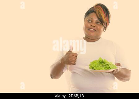 Femme africaine noire grasse mûrie et attentionnés souriant et donnant le pouce tout en tenant la laitue servie sur une assiette blanche Banque D'Images