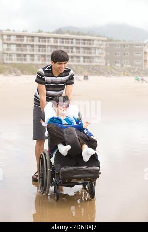 Jeune biracial asiatique Caucasien poussant le petit frère handicapé dedans fauteuil roulant sur la plage Banque D'Images