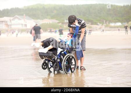 Jeune biracial asiatique Caucasien poussant le petit frère handicapé dedans fauteuil roulant sur la plage Banque D'Images