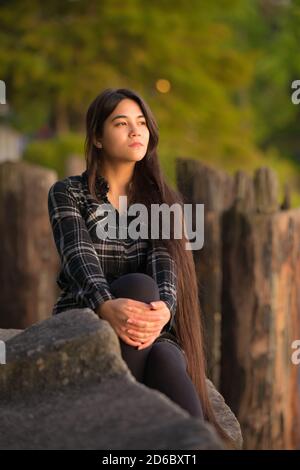 Jeune femme asiatique biraciale de race blanche assise seule sur des rochers à côté jetée en bois donnant sur le coucher du soleil Banque D'Images