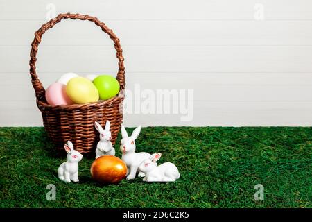Joyeuses Pâques. Félicitations de pâques. Œufs de Pâques colorés dans un panier avec des petits lapins blancs autour de l'œuf doré sur un fond de mousse et un mur en bois clair avec espace de copie. Banque D'Images