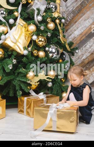 Portrait de jeune fille d'âge élémentaire portant robe bleue assise à côté de grand déballage de l'arbre de Noël cadeau enveloppé fort curieux à propos d'un présent le lendemain de Banque D'Images