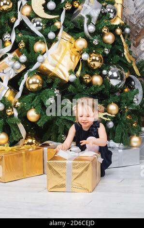 Portrait de jeune fille d'âge élémentaire portant robe bleue assise à côté de grand déballage de l'arbre de Noël cadeau enveloppé fort curieux à propos d'un présent le lendemain de Banque D'Images